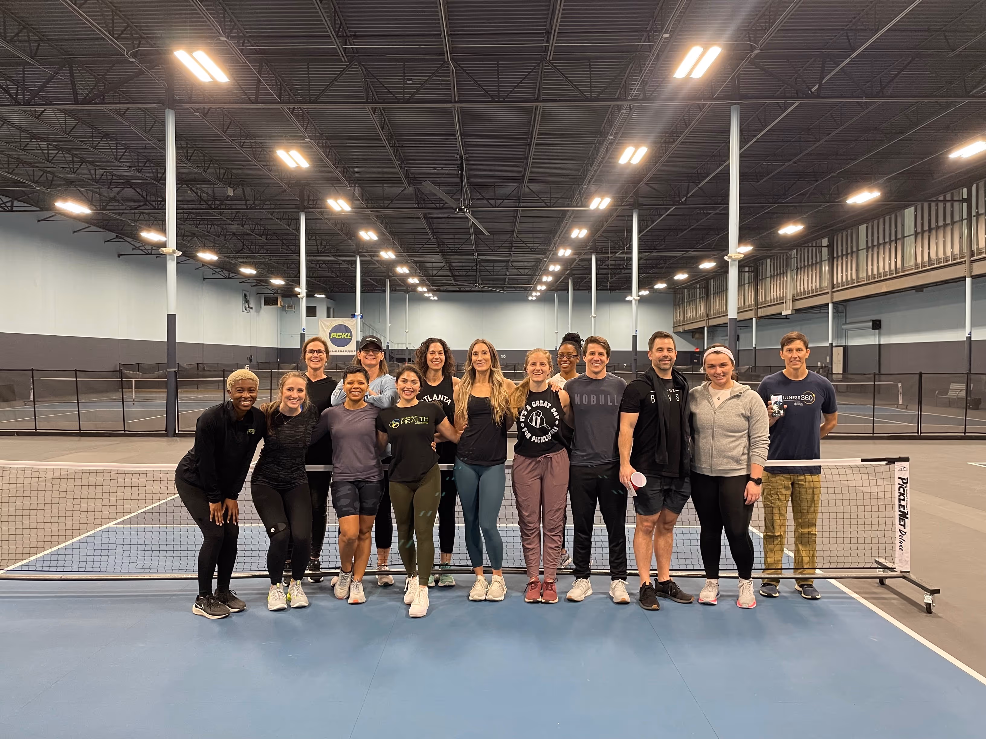 Group of fifteen people posing and smiling together on an indoor pickleball court behind the net.