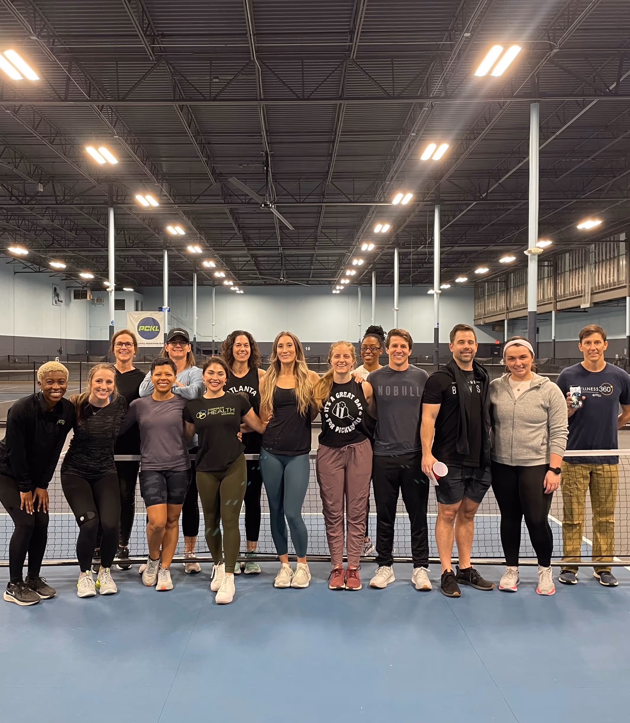 Group of fifteen people standing on an indoor pickleball court posing for a photo behind the net.