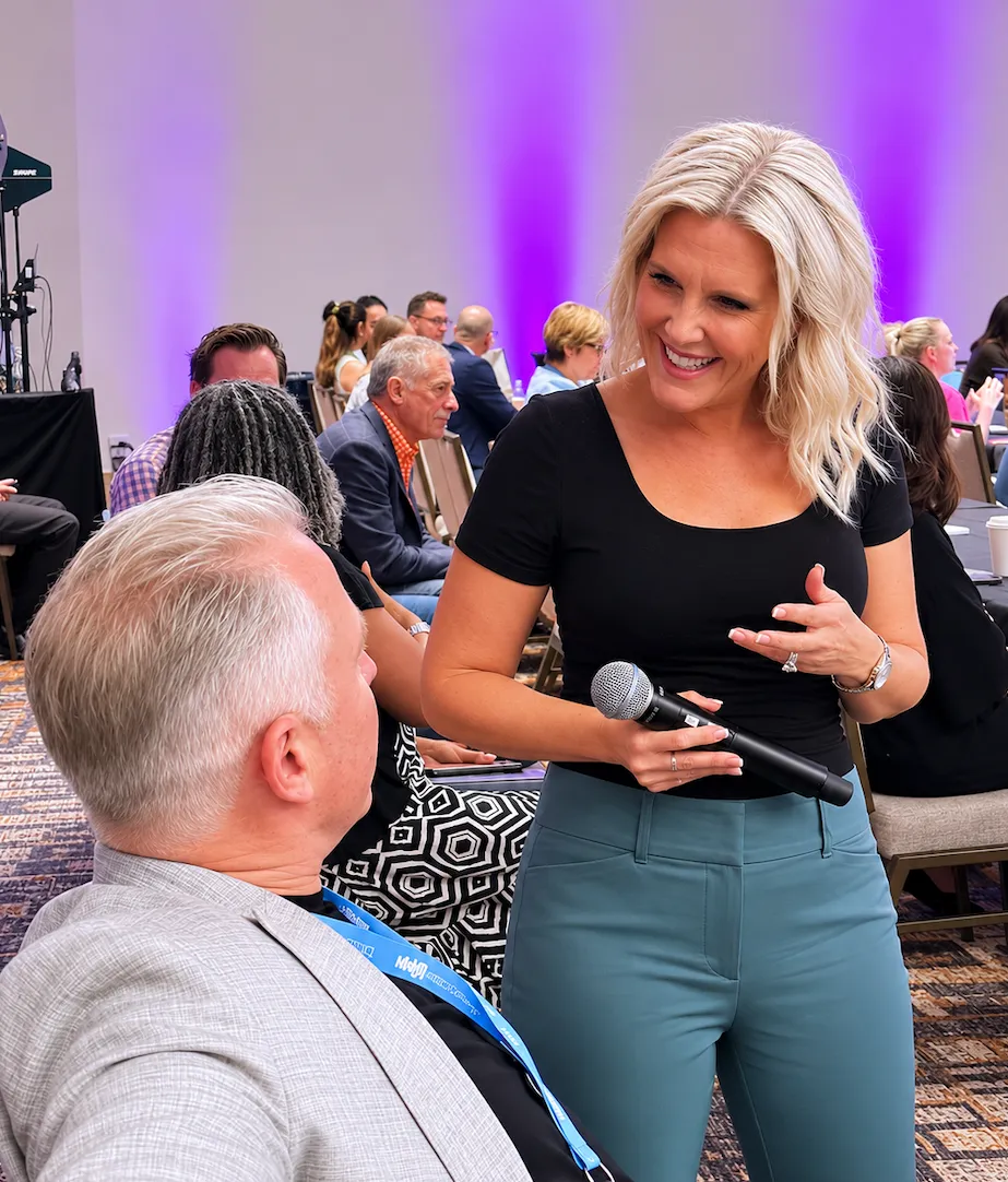 Woman with microphone smiling and talking to seated man in a conference room with purple lighting.