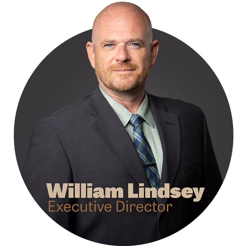 Portrait of William (Billy) Lindsey from DeKalb, TX, in a dark suit, green shirt, and blue plaid tie against a dark background.