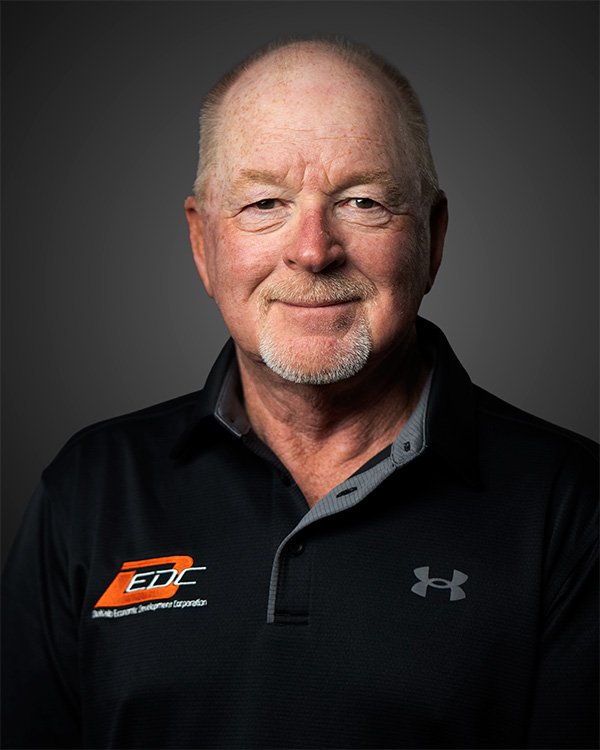 Portrait of a smiling older man with short gray hair and goatee wearing a black polo shirt.