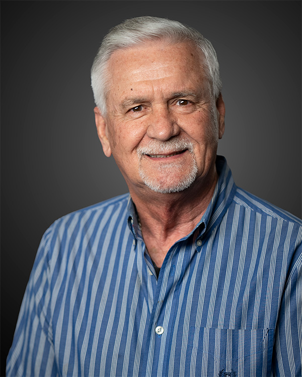 Portrait of a smiling older man with white hair wearing a blue striped shirt against a dark background.