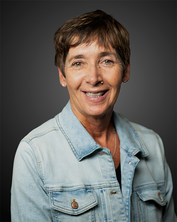 Smiling middle-aged woman with short brown hair wearing a light denim jacket against a gray background.