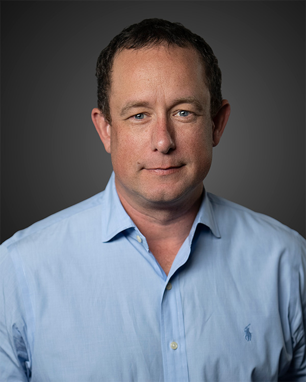 Man with short curly hair wearing a light blue collared shirt against a gray background.