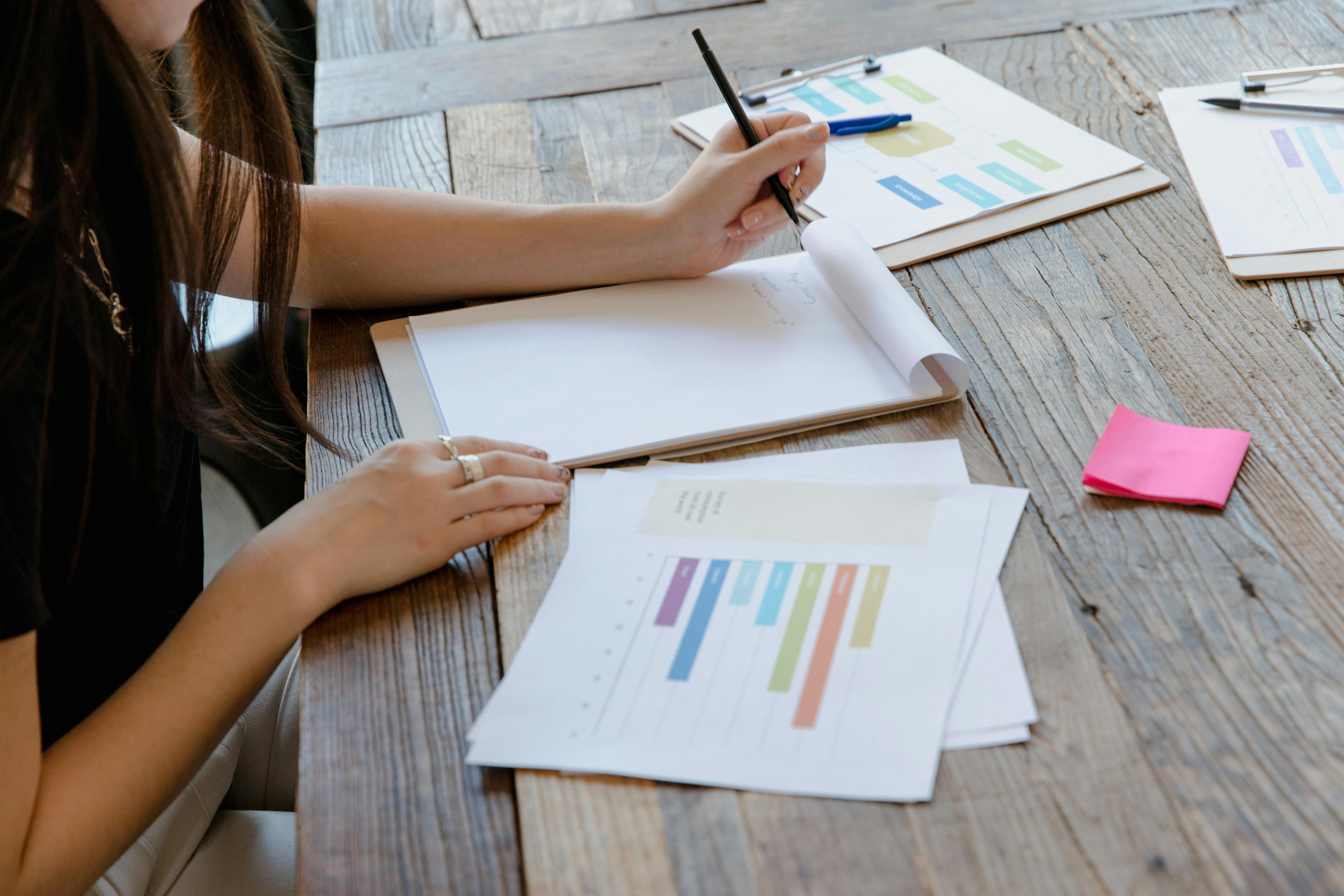 Person writing on a notepad at a wooden table with charts and colorful sticky notes.