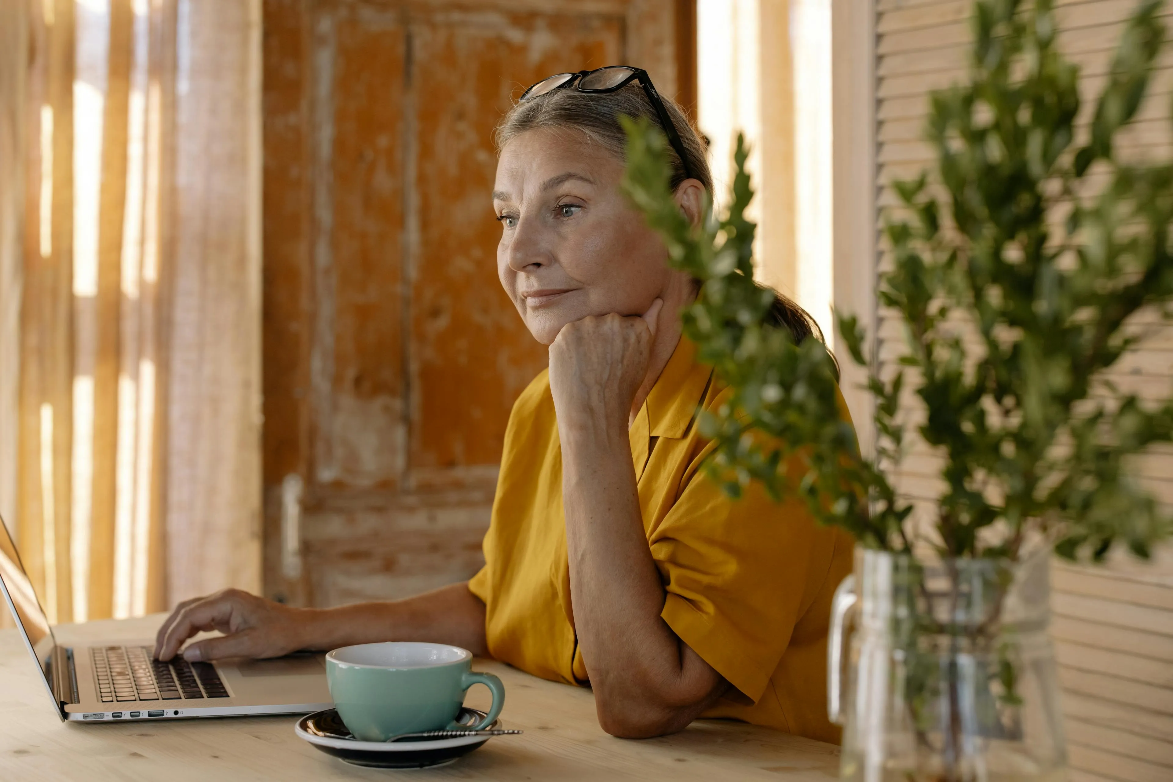 Woman in a yellow shirt using a laptop at a wooden table with a teal coffee cup and green plant in a glass vase.