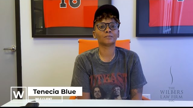 Person wearing a baseball cap and glasses sitting in an orange chair in front of framed sports jerseys, identified as Tenecia Blue, WLF client.