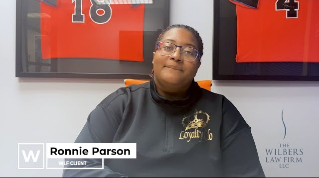 A woman wearing glasses and a black Loyalty Co sweatshirt sitting in a chair with framed sports jerseys on the wall behind her, identified as Ronnie Parson, WLF client.