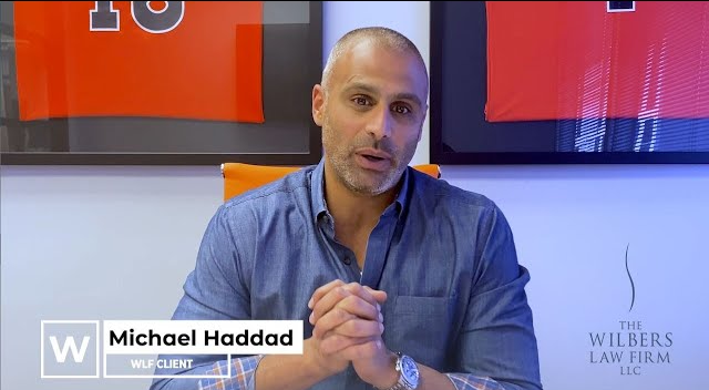 Man in a blue shirt sitting with hands clasped in front of framed orange sports jerseys, labeled as Michael Haddad, WLF client, with The Wilbers Law Firm logo.