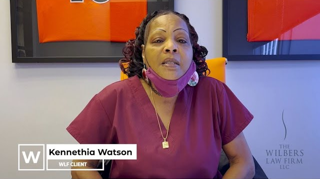 Kennethia Watson, a WLF client, sitting in an office wearing a maroon scrub top and a purple face mask under her chin.