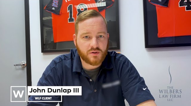 Man with red hair and beard wearing a navy blue shirt, sitting in an office with framed sports jerseys on the wall behind him, and a Wilbers Law Firm logo on the right.
