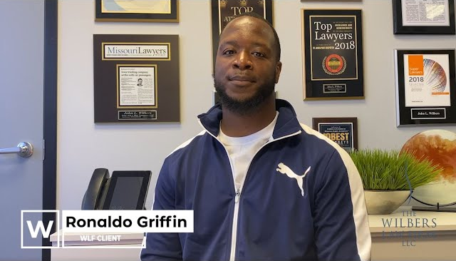 Man in a blue and white Puma jacket sitting in an office with framed awards and certificates on the wall behind him.