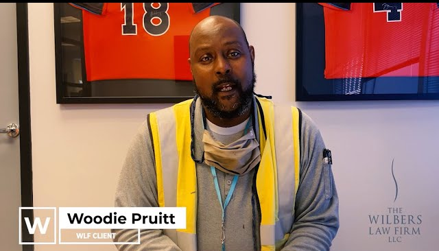 Woodie Pruitt, wearing a yellow safety vest and grey shirt, seated indoors with framed sports jerseys on the wall behind him.