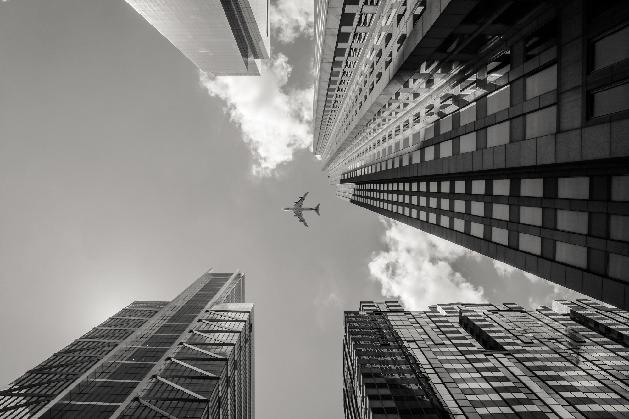 Airplane flying above tall skyscrapers against a partly cloudy sky, viewed from the ground.