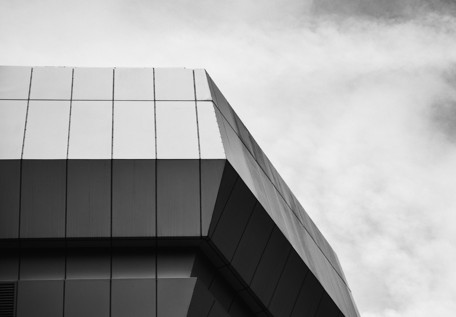 Angular, modern building facade with geometric panels against a cloudy sky in black and white.