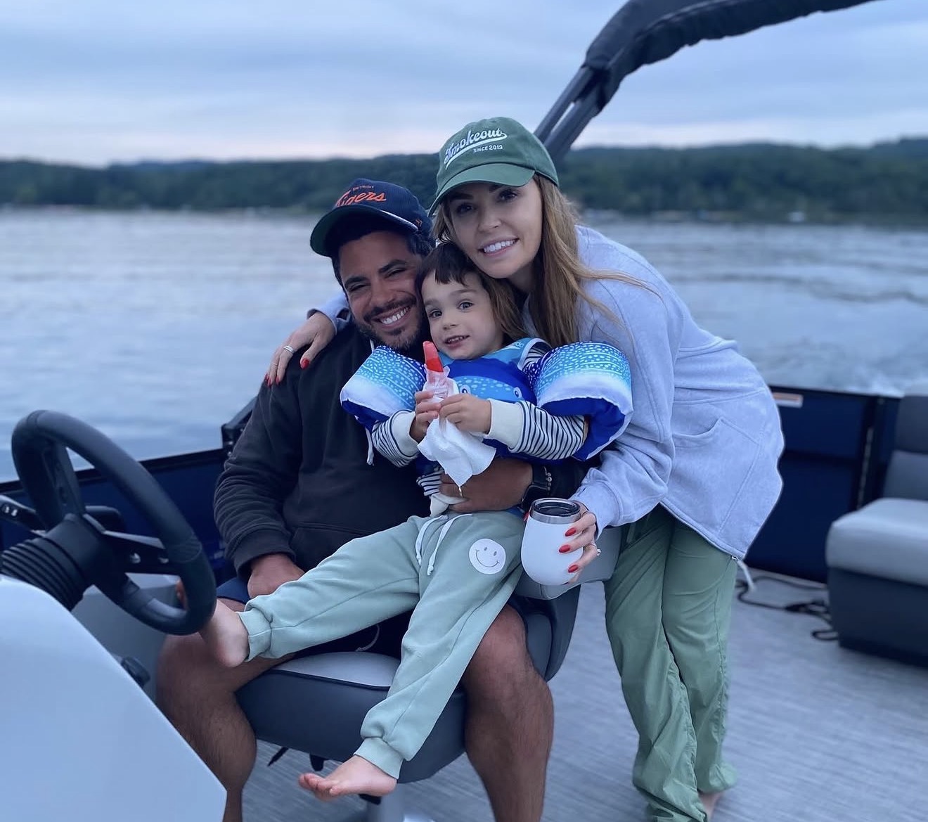 A smiling family of three on a boat with water and trees in the background during dusk.