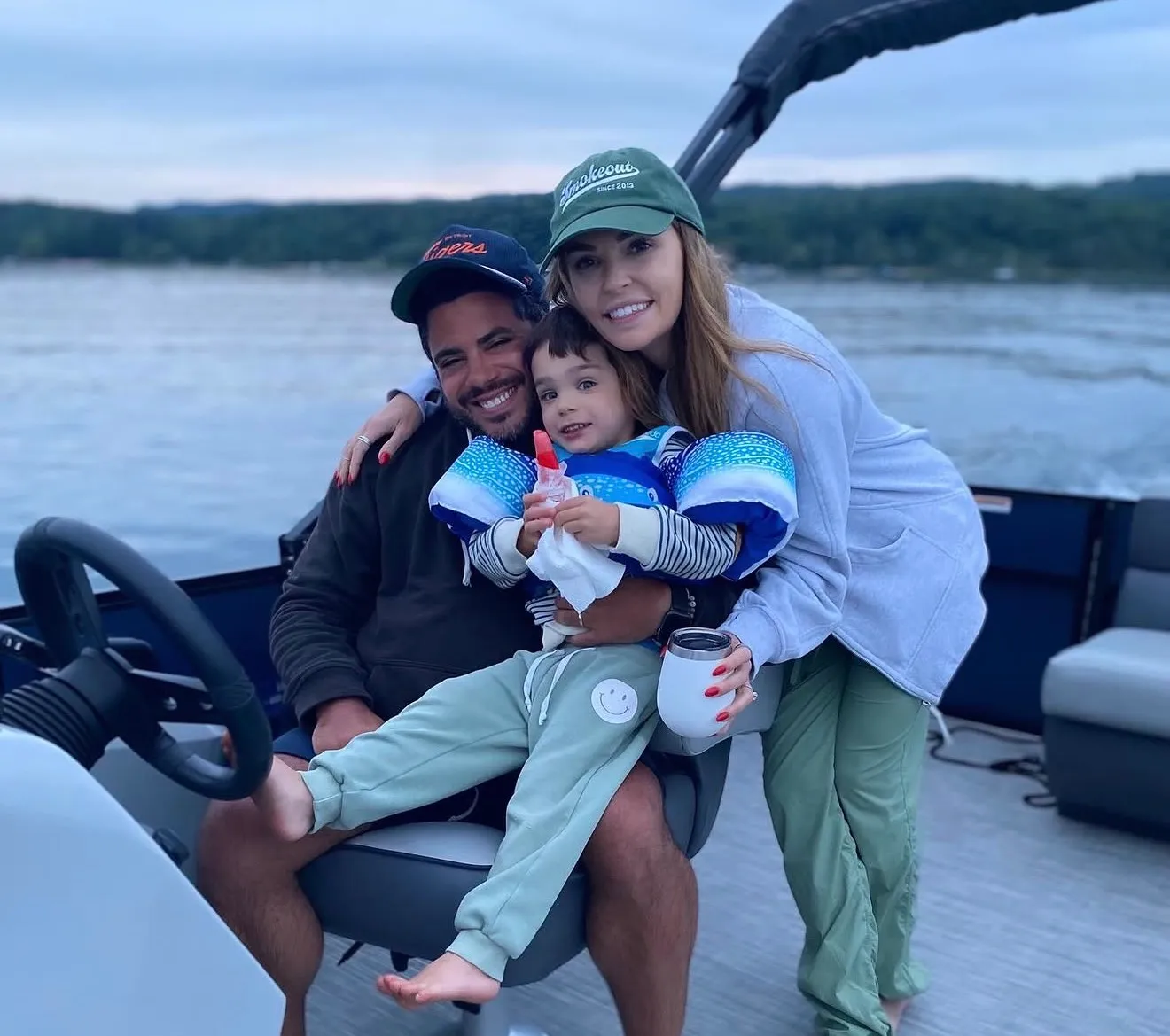 A smiling family of three on a boat with water and trees in the background during dusk.