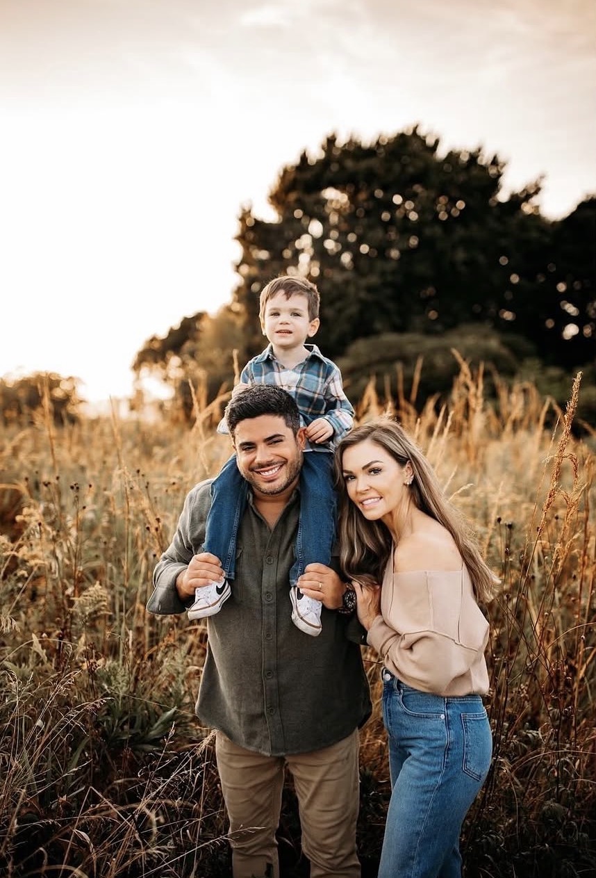 Smiling family outdoors with father carrying young son on shoulders and mother standing beside them.