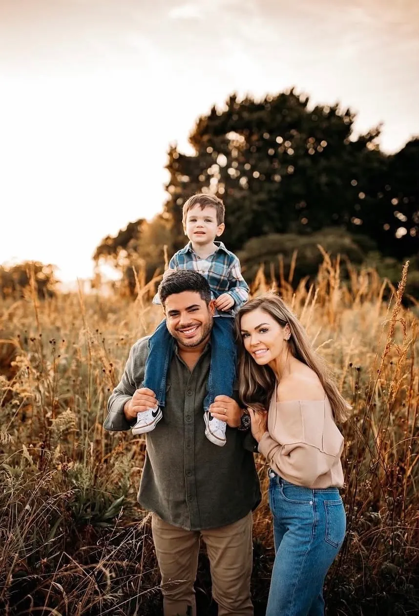 Smiling family outdoors with father carrying young son on shoulders and mother standing beside them.