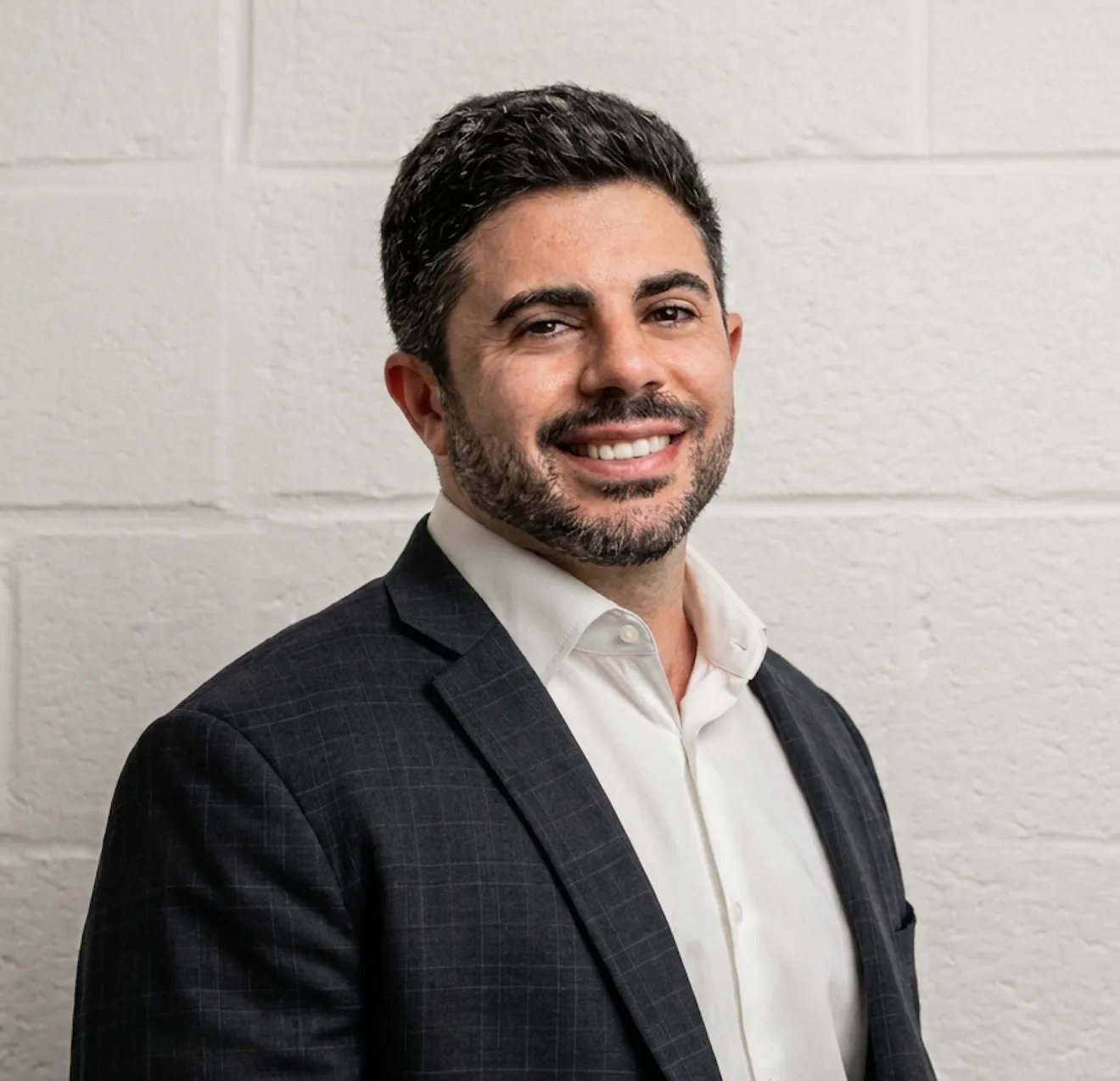 Smiling man with short dark hair and beard wearing a white shirt and dark checkered suit jacket.
