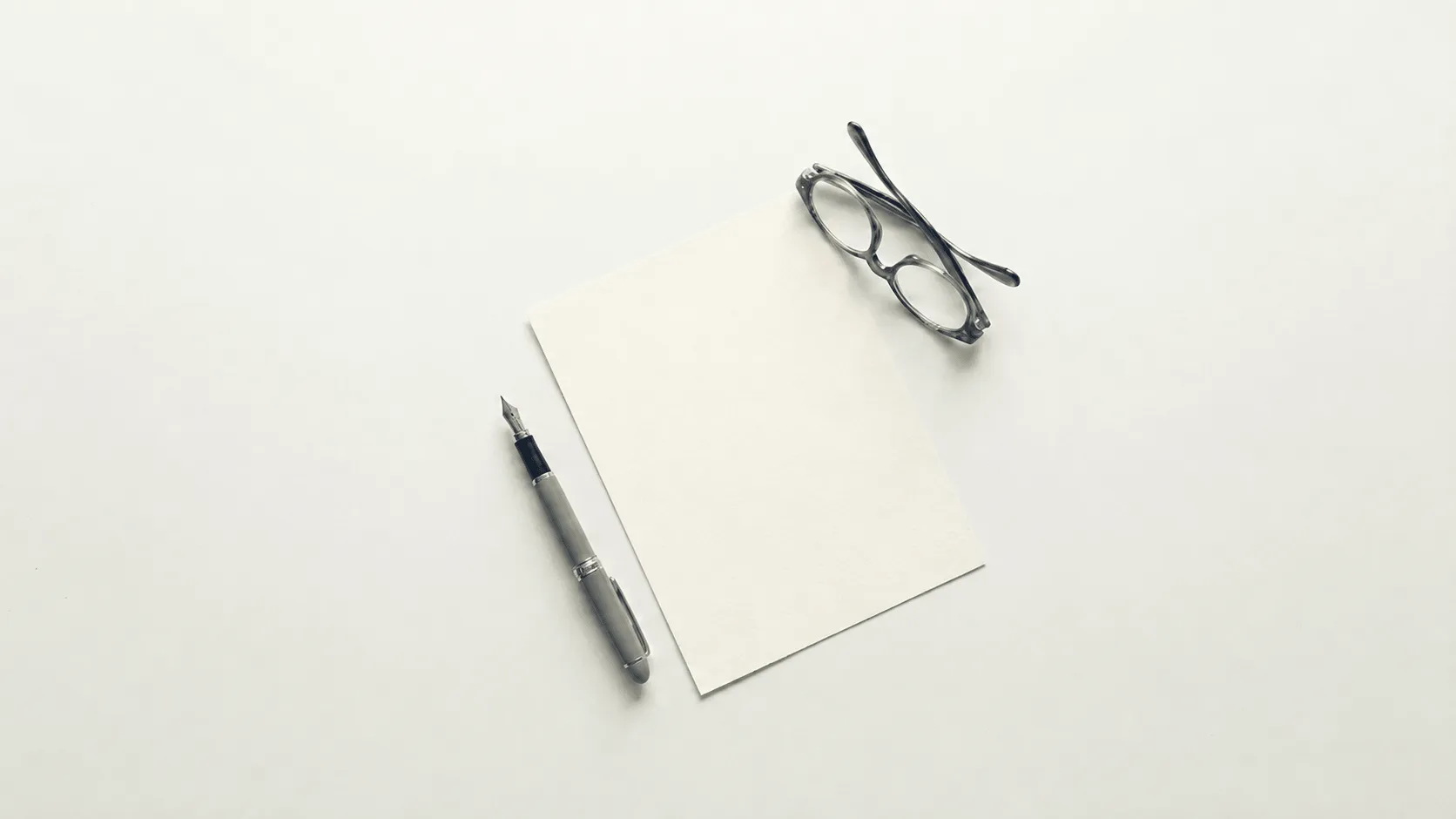 Blank sheet of paper with a fountain pen and eyeglasses on a white surface.