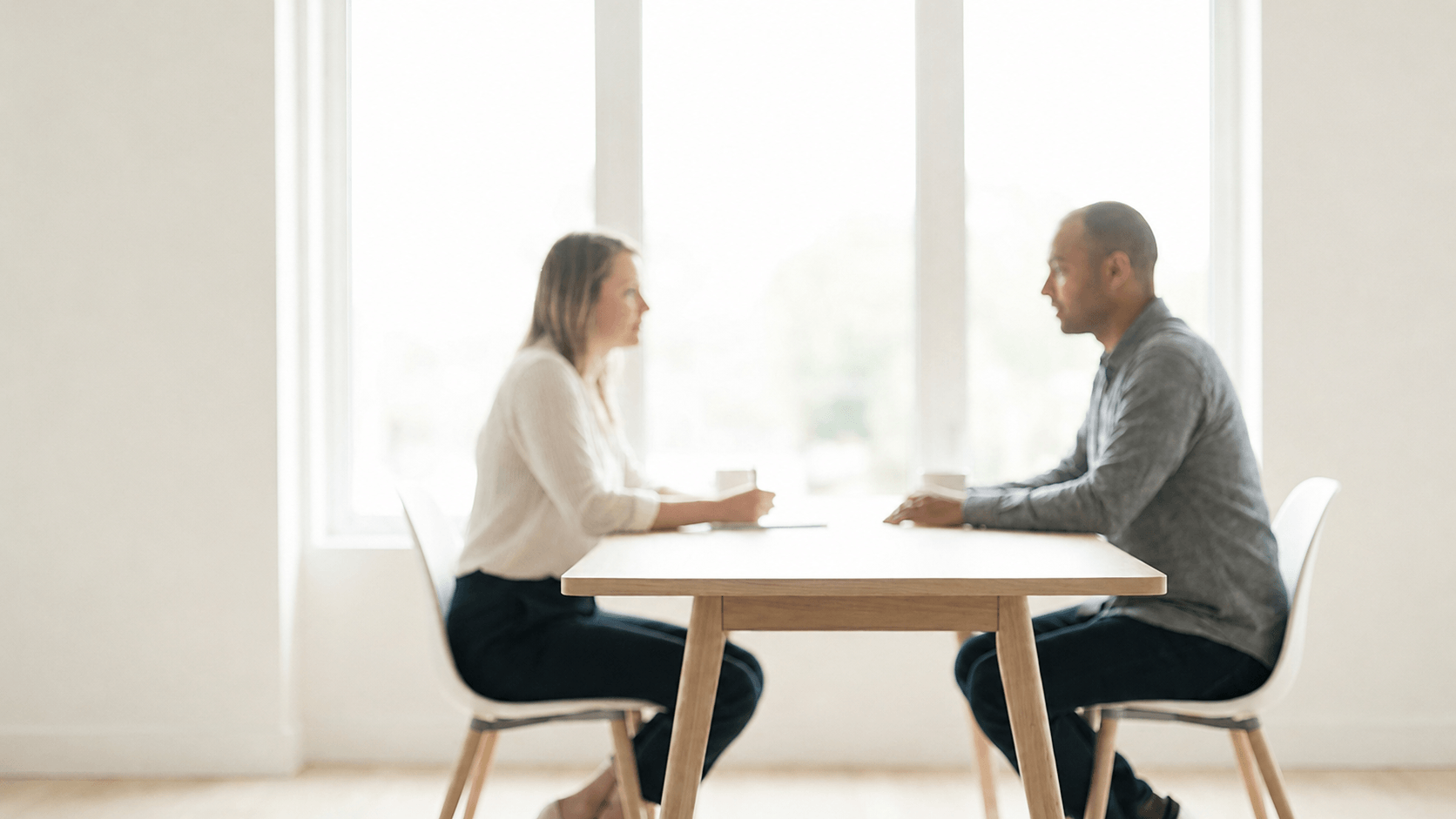 A man and woman sit across from each other at a wooden table in a bright room engaged in conversation.