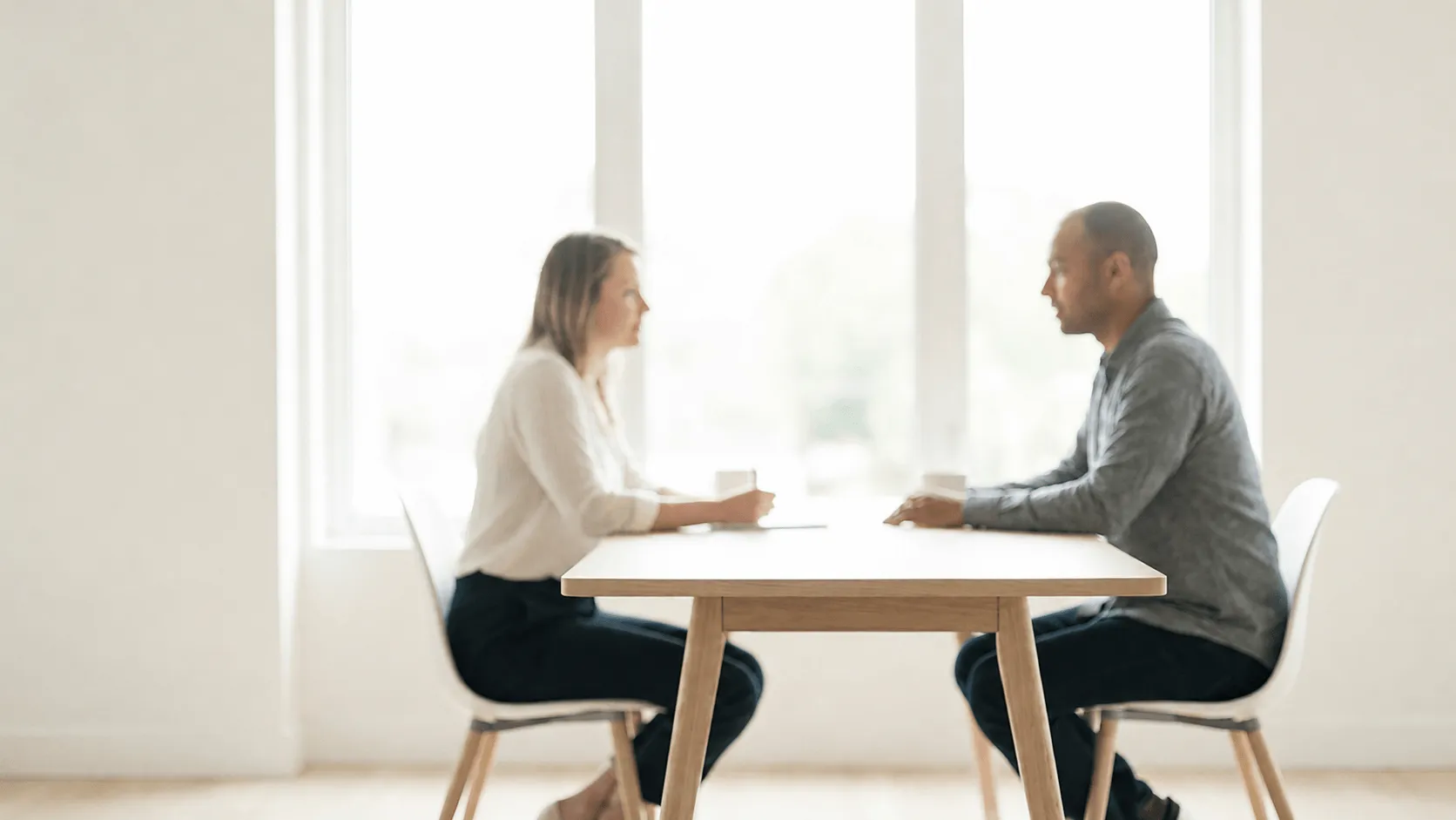 A man and woman sit across from each other at a wooden table in a bright room engaged in conversation.