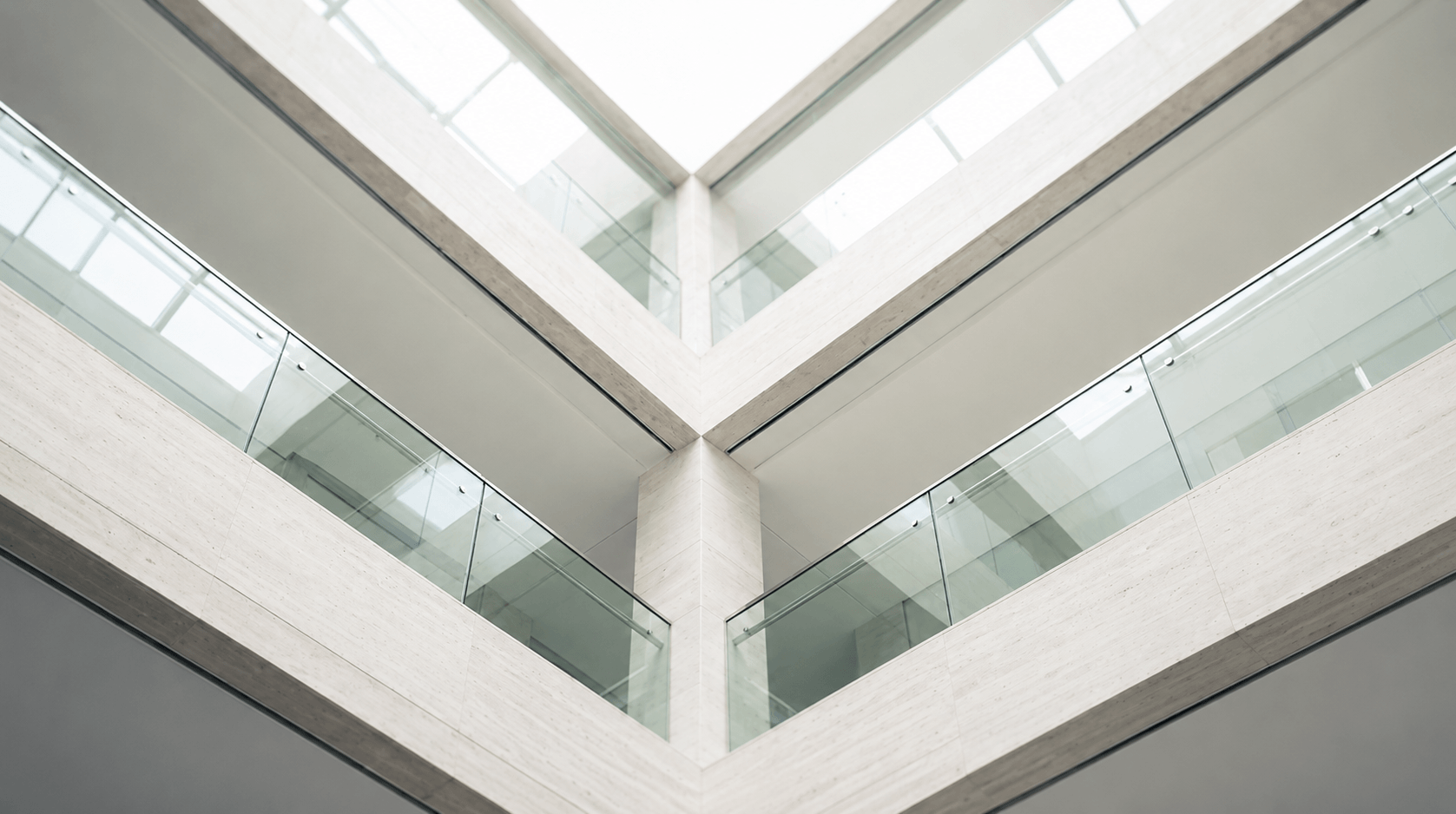 Looking up at a modern building corner with glass railings and pale stone walls under a skylight.