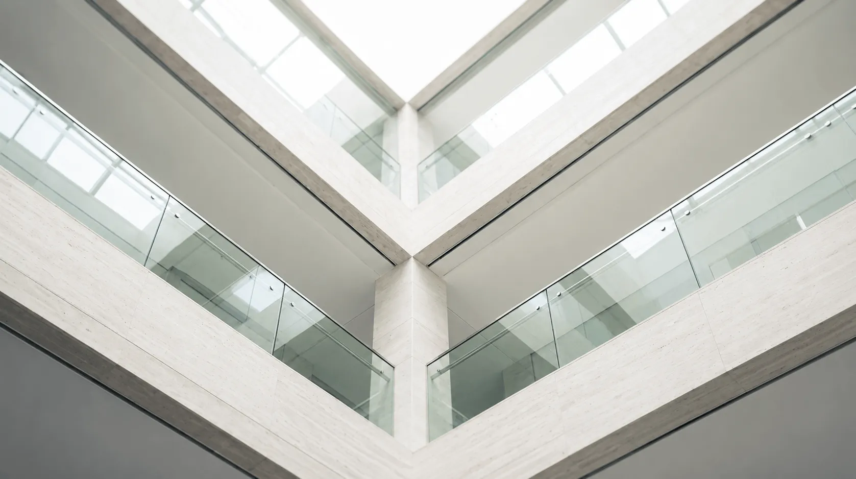 Looking up at a modern building corner with glass railings and pale stone walls under a skylight.