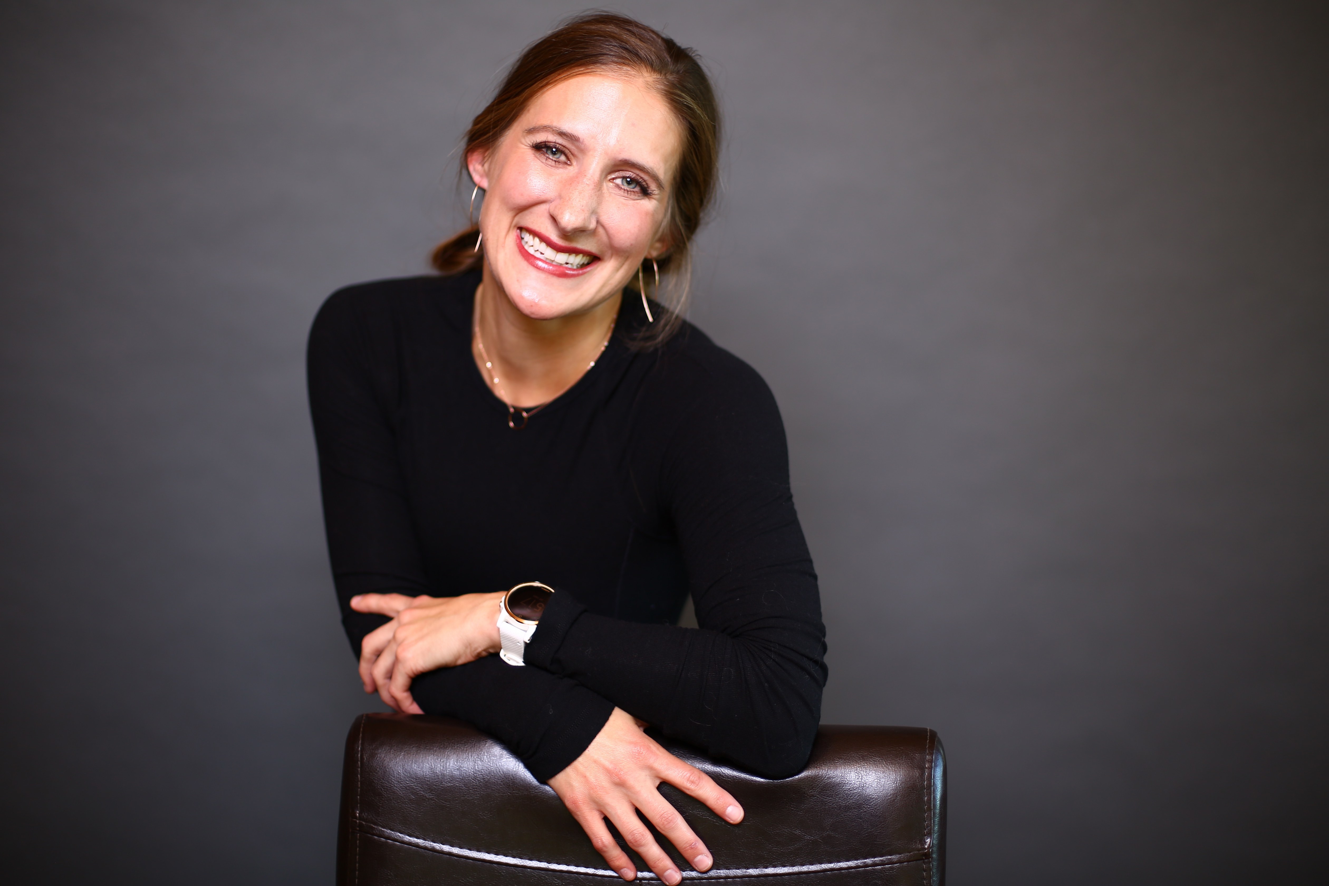 Smiling woman in black long sleeve leans on chair back against gray background.