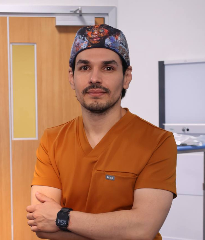 Man in orange scrubs and printed surgical cap standing with arms crossed in a medical setting.