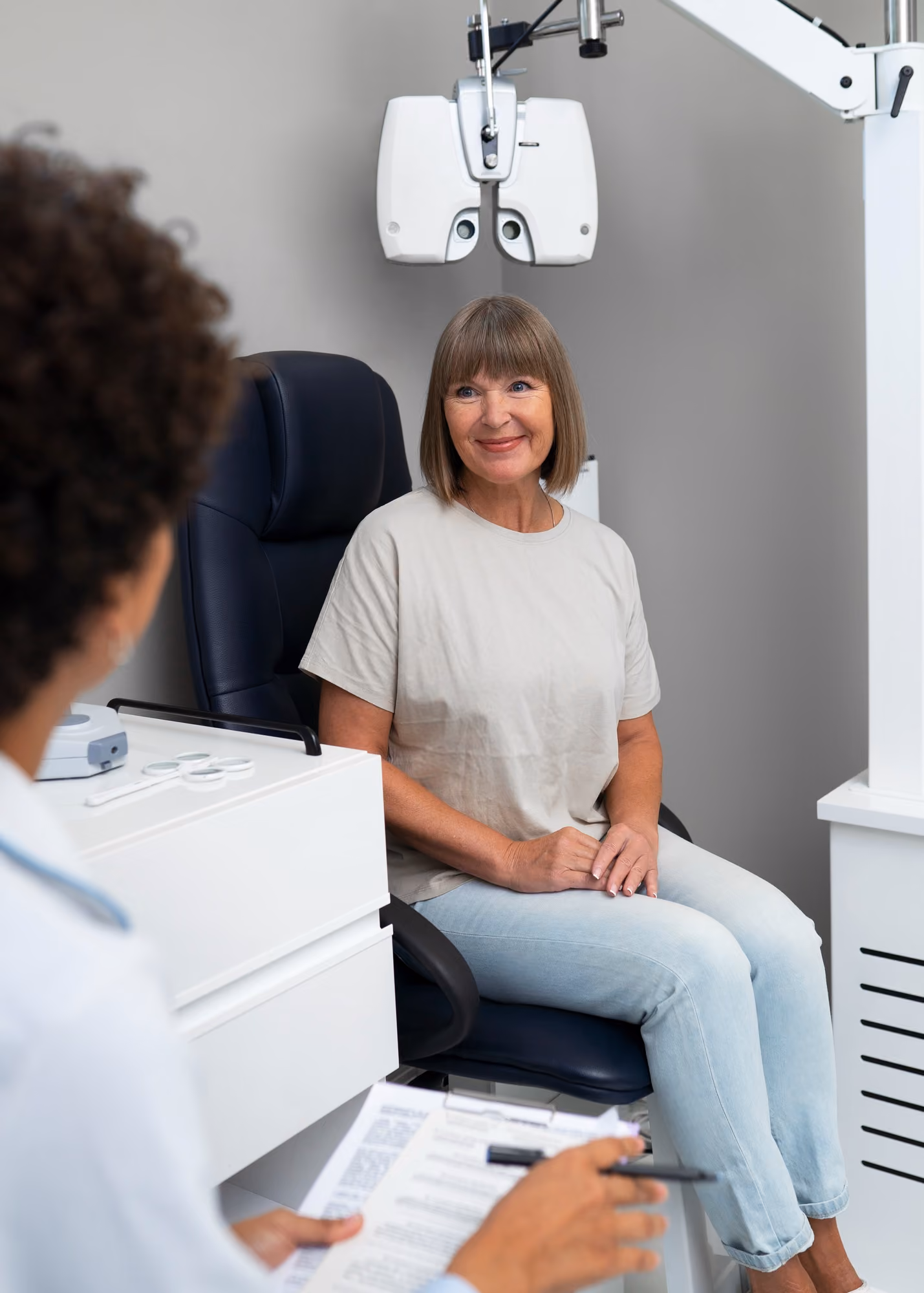 Smiling mature woman sitting in an eye exam chair with an optometrist holding a clipboard nearby.