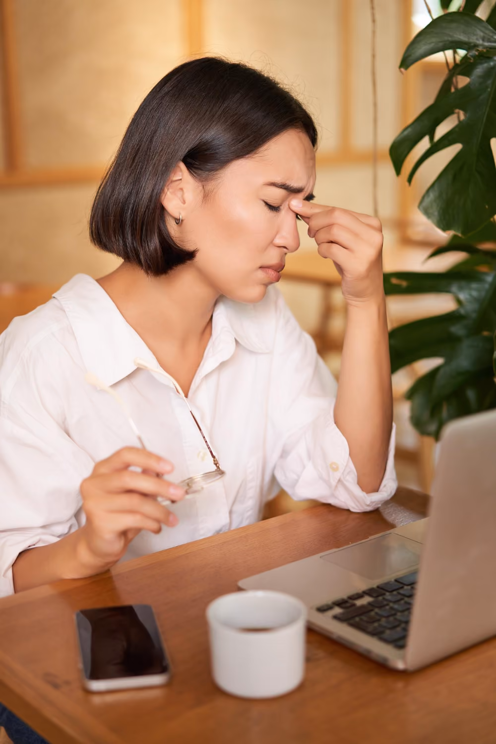 Woman sitting at a table with laptop, holding glasses and rubbing her eyes, appearing stressed or tired.