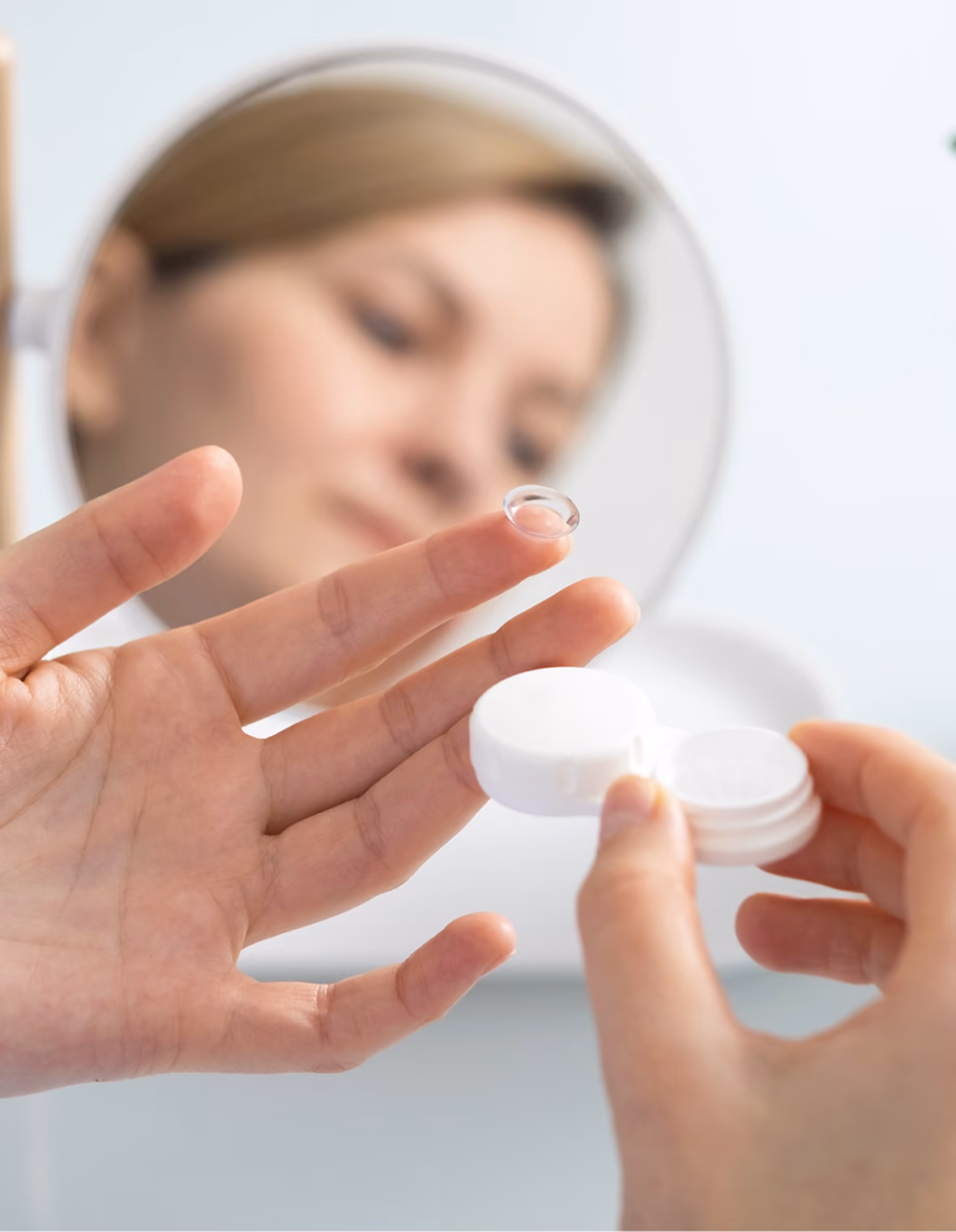 A hand holding a clear contact lens on a finger with a white lens case nearby and a blurred face in the mirror.