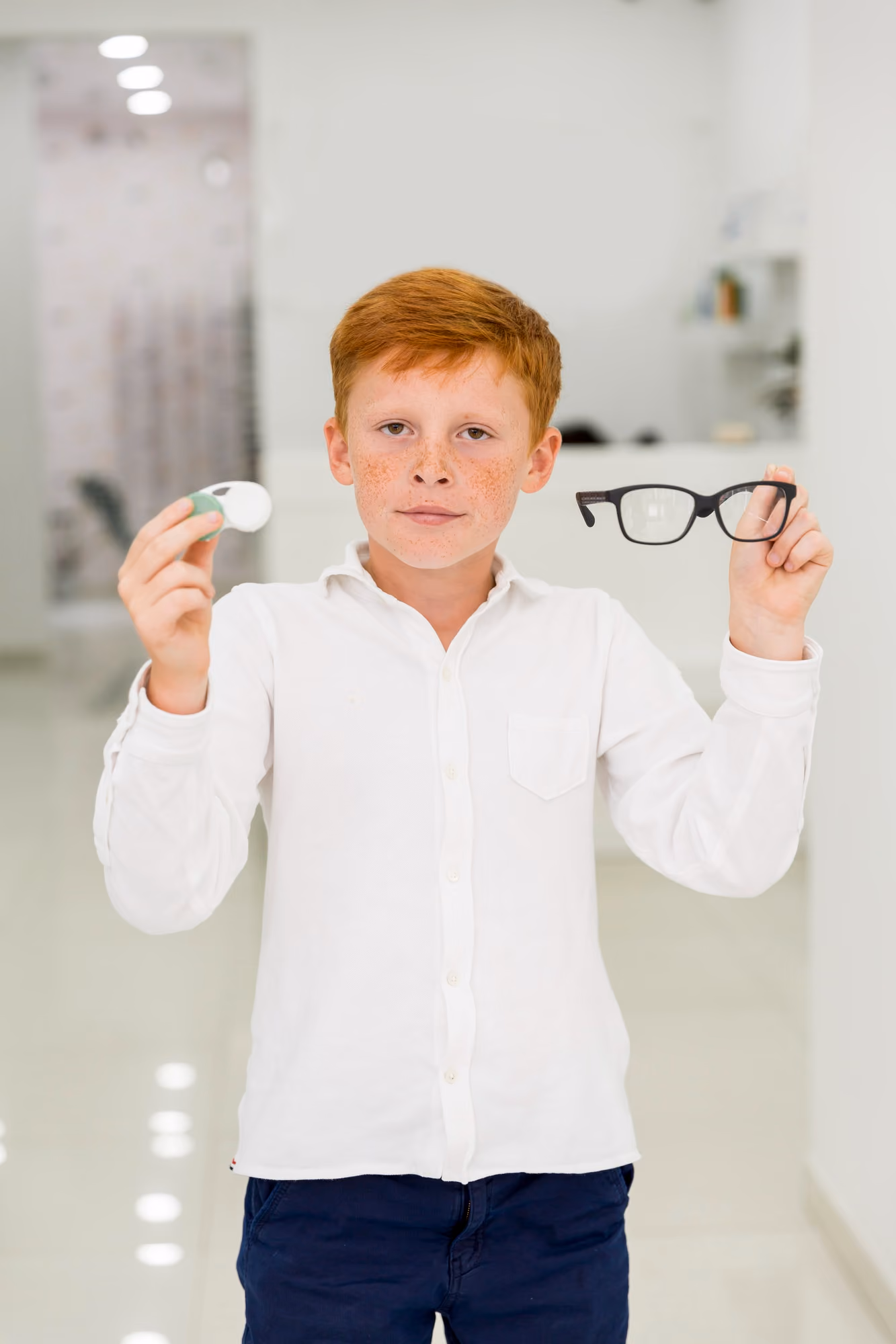 Young boy with red hair and freckles holding black eyeglasses in one hand and contact lens case in the other.