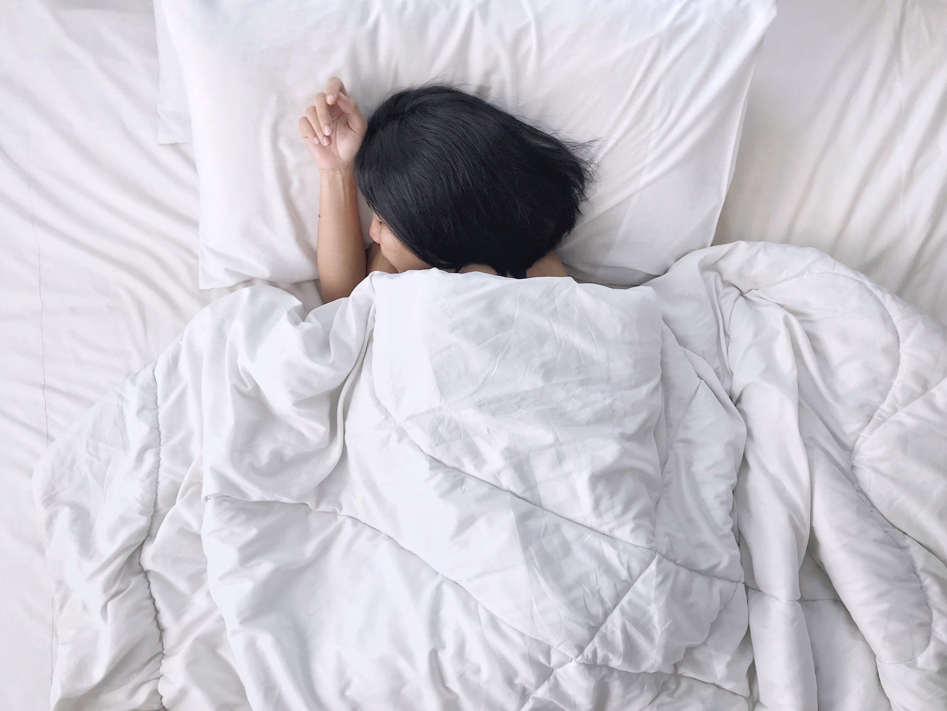 Person with black hair sleeping on white bed under white blanket with white pillow.