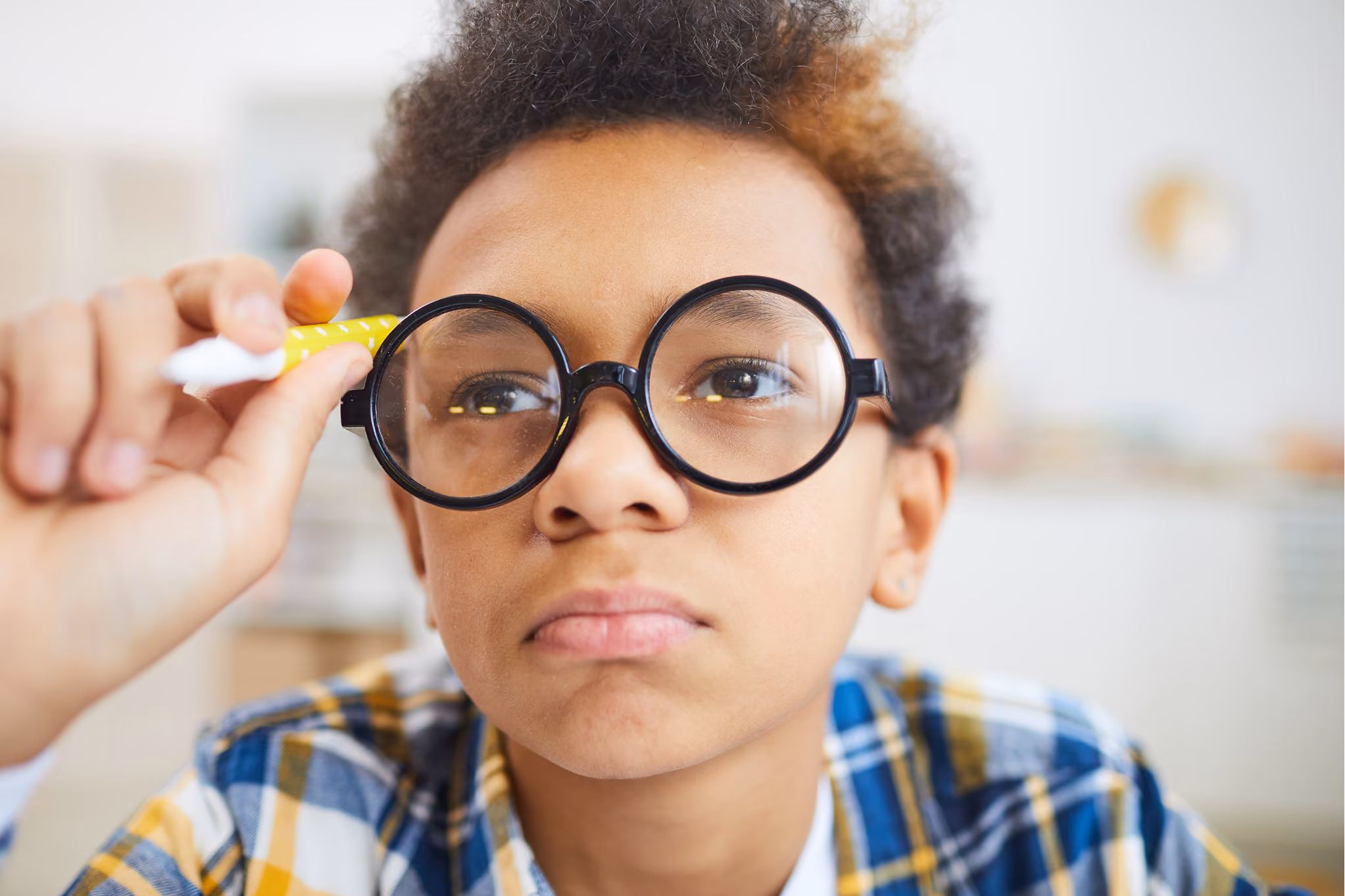 Boy with curly hair wearing large round glasses holds pen while looking intently ahead.