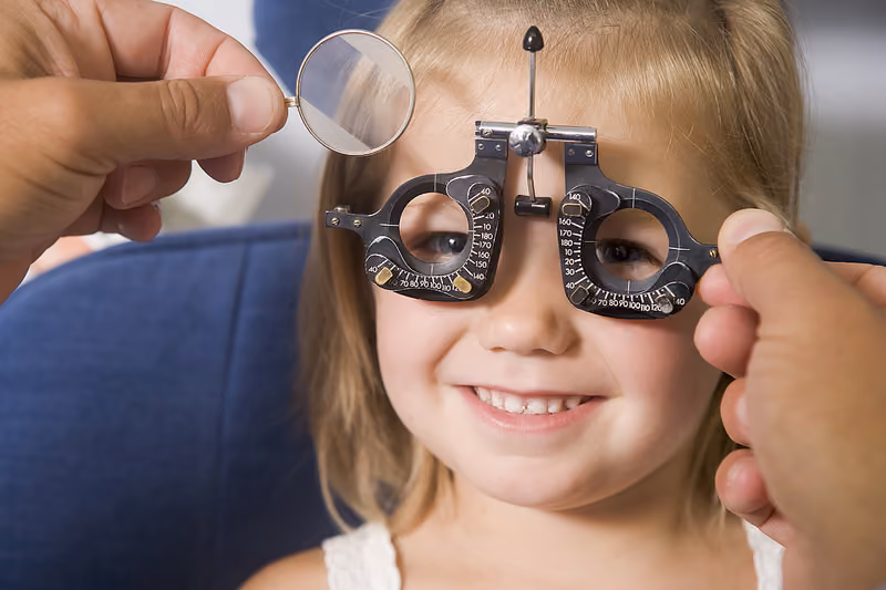 A young girl smiling during an eye exam with a trial frame and lens held by an optometrist.