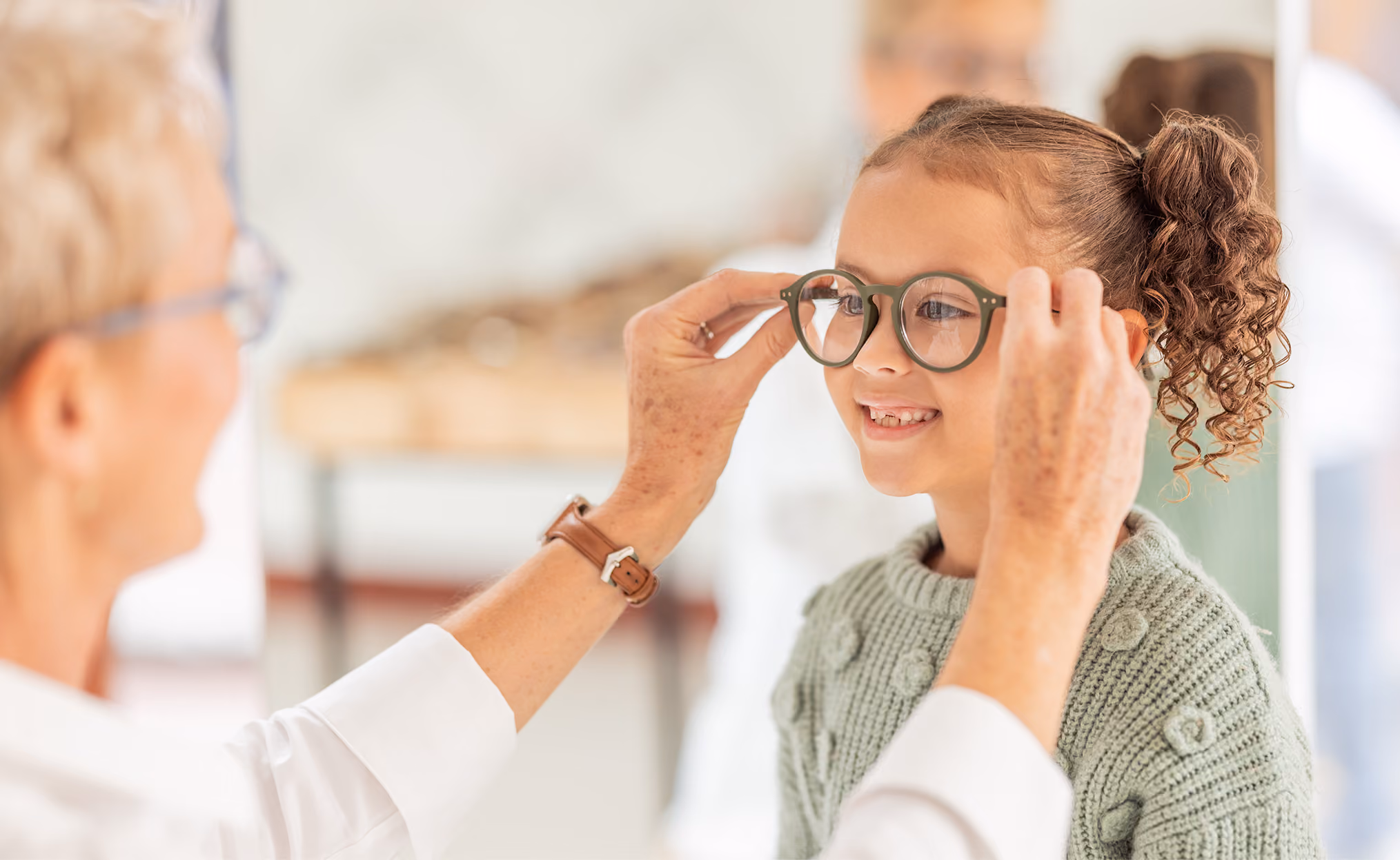 An older woman fitting eyeglasses on a smiling young girl with curly hair in a bun.