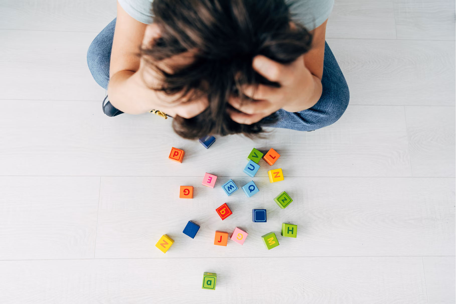 Top view of a child sitting on floor with head in hands, scattered colorful letter blocks nearby.