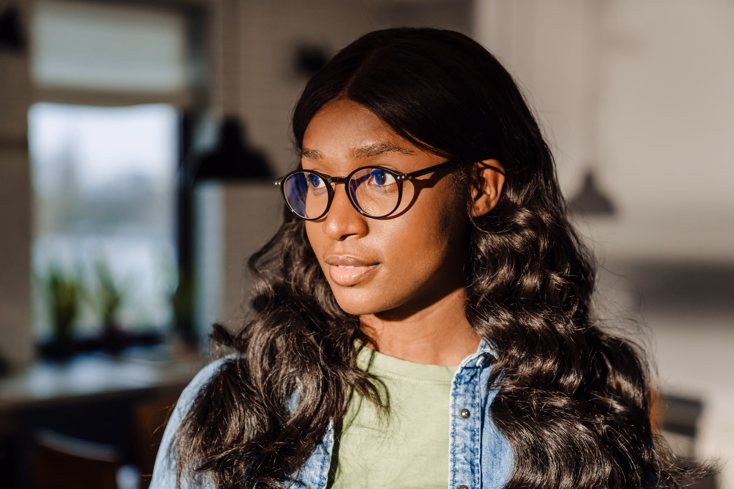 Young woman with long wavy hair wearing glasses and a denim jacket looking to the side indoors.