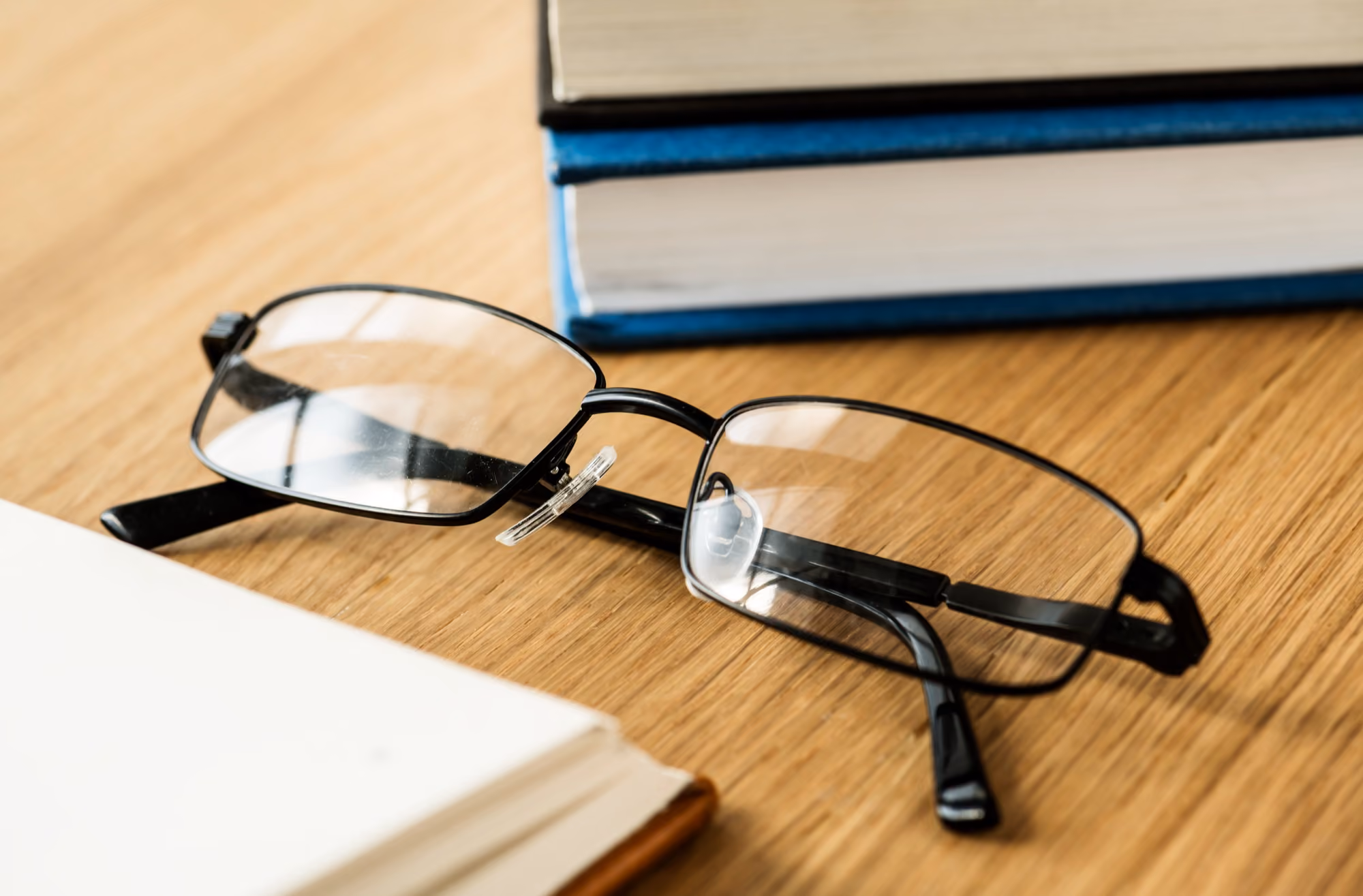 Black-framed eyeglasses lying on a wooden table near a stack of books and an open book.