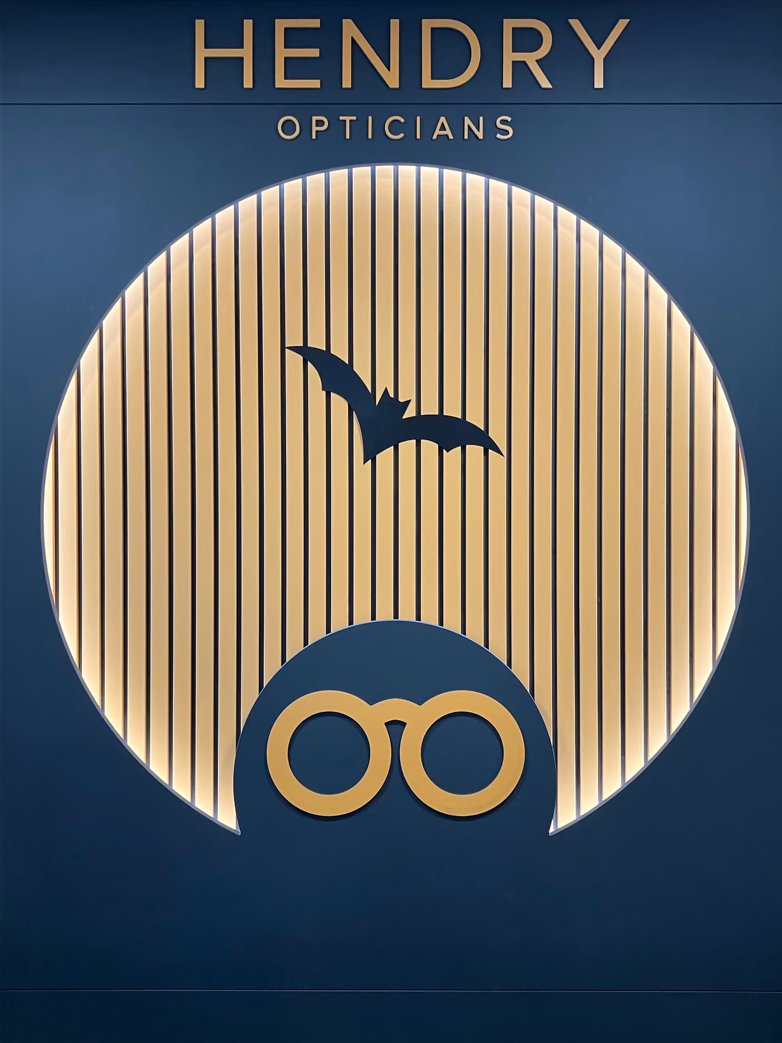 HENDRY OPTICIANS sign with bat silhouette and round eyeglasses icon on dark blue background.