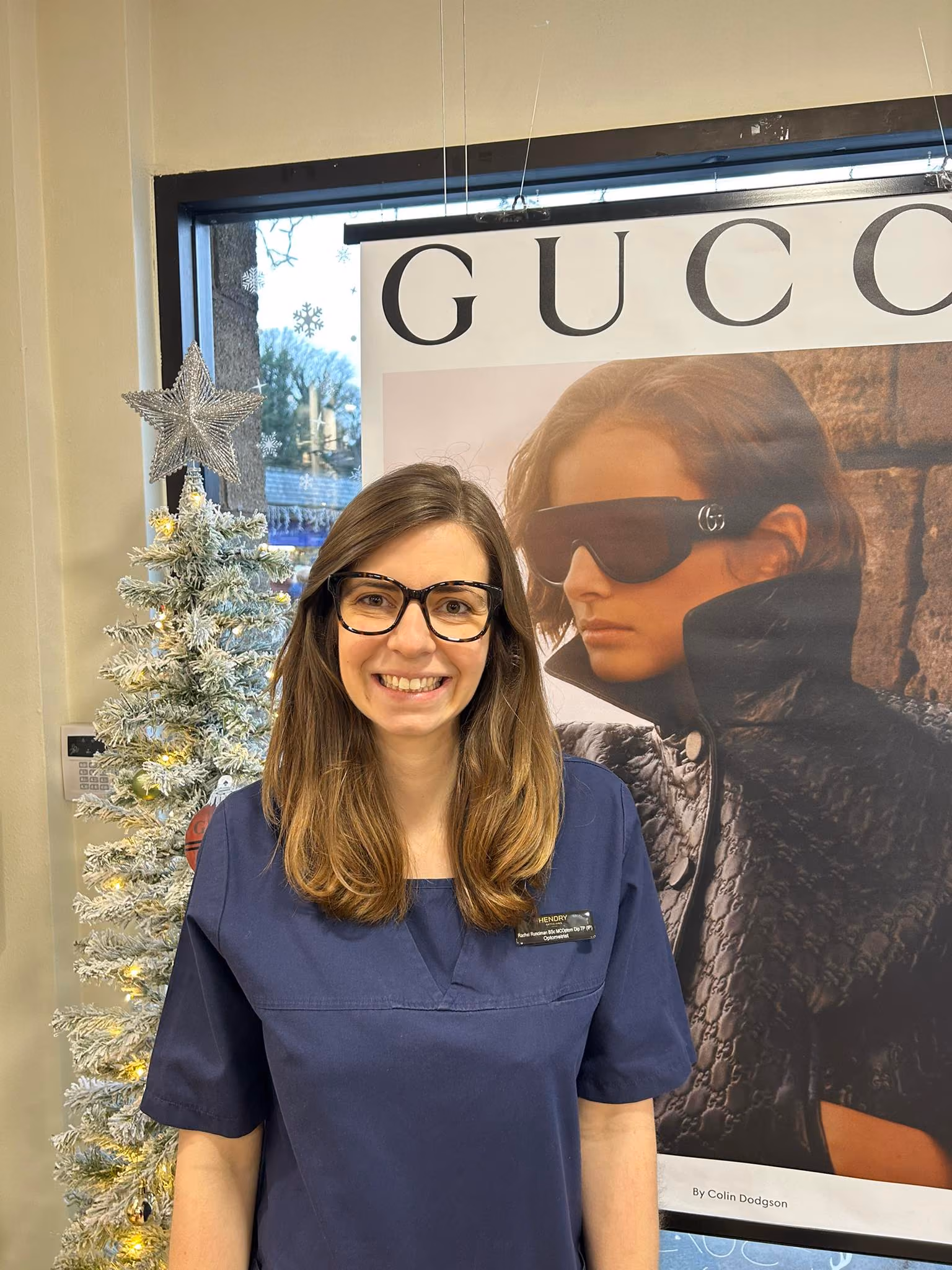 Smiling woman with glasses in navy blue scrubs stands next to a decorated white Christmas tree and Gucci poster.