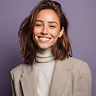Smiling young woman with wavy brown hair wearing a beige blazer and white turtleneck against a purple background.