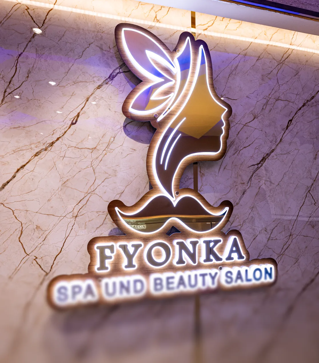 Leuchtendes Logo von Fyonka Spa und Schönheitssalon mit Silhouette eines Frauengesichts und Schmetterlingsflügeln an einer Marmorwand.