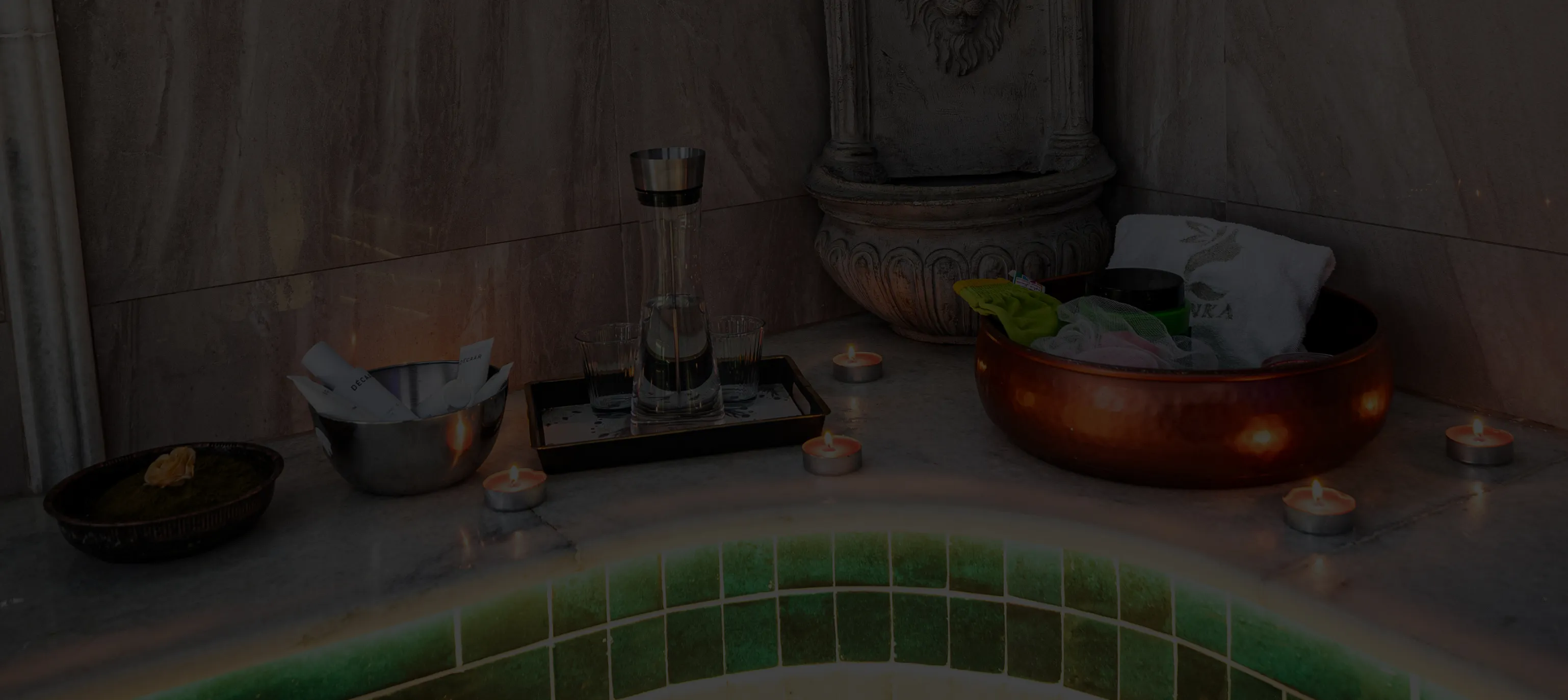 Beleuchtetes Spa-Bad mit grün gekacheltem Rand, Teelichtern, Karaffe mit Wasser, Gläsern und Pflegeutensilien auf Marmorablage.