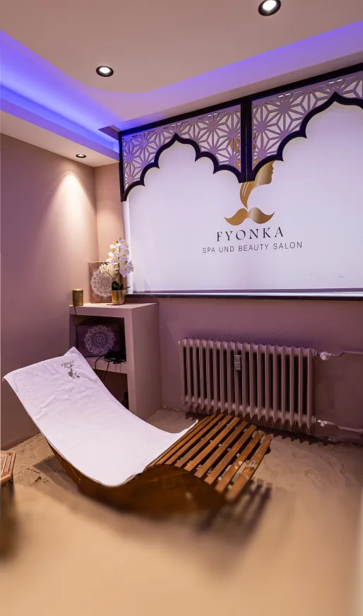 Entspannender Spa-Raum mit ergonomischem Holzliegestuhl, weißen Kissen, Orchideen und einem beleuchteten Fyonka Spa und Beauty Salon-Logo an der Wand.