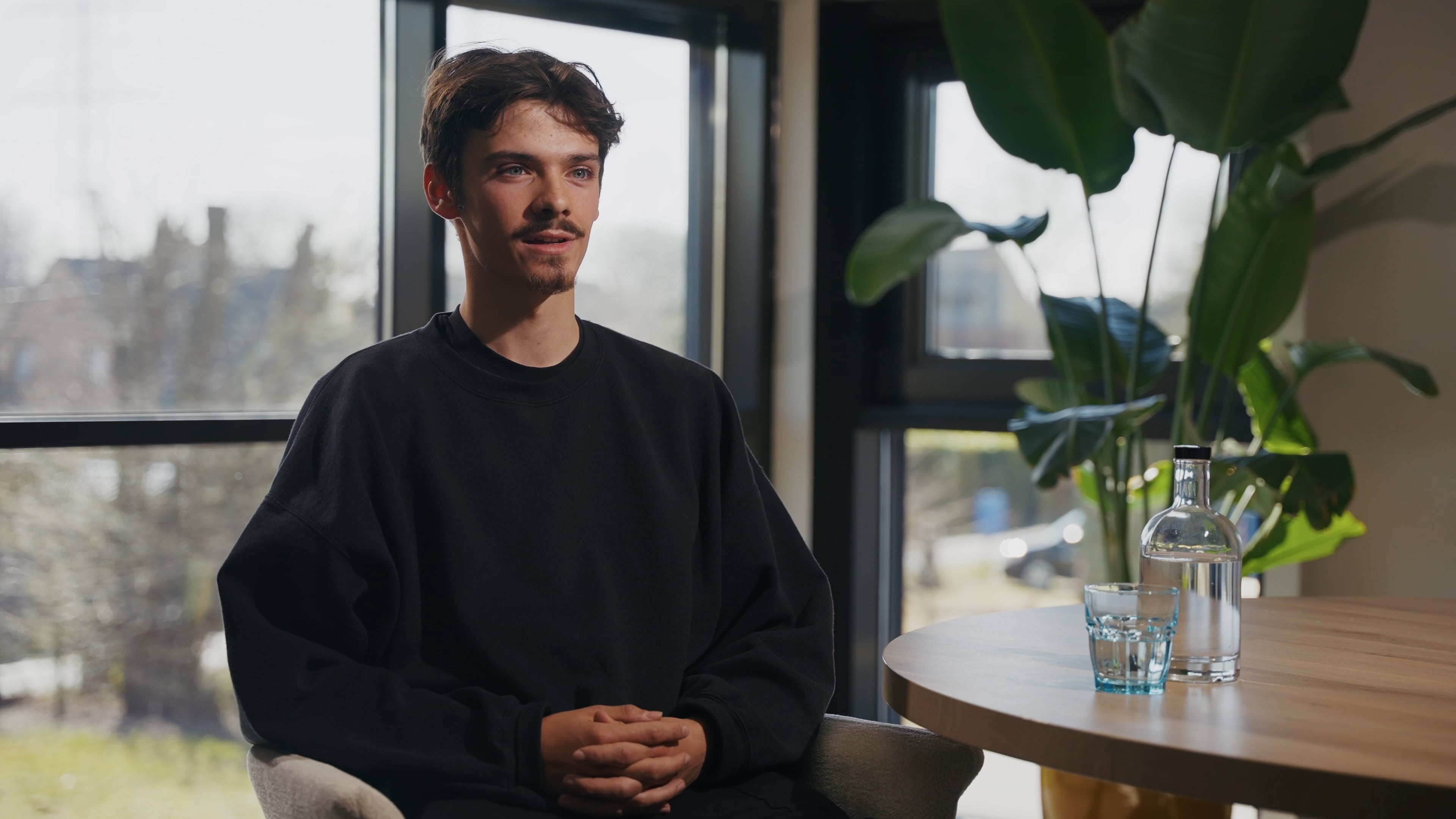 Een jonge man met kort donker haar en een snor, zittend aan een tafel bij het raam, met een glas en een fles water op tafel.