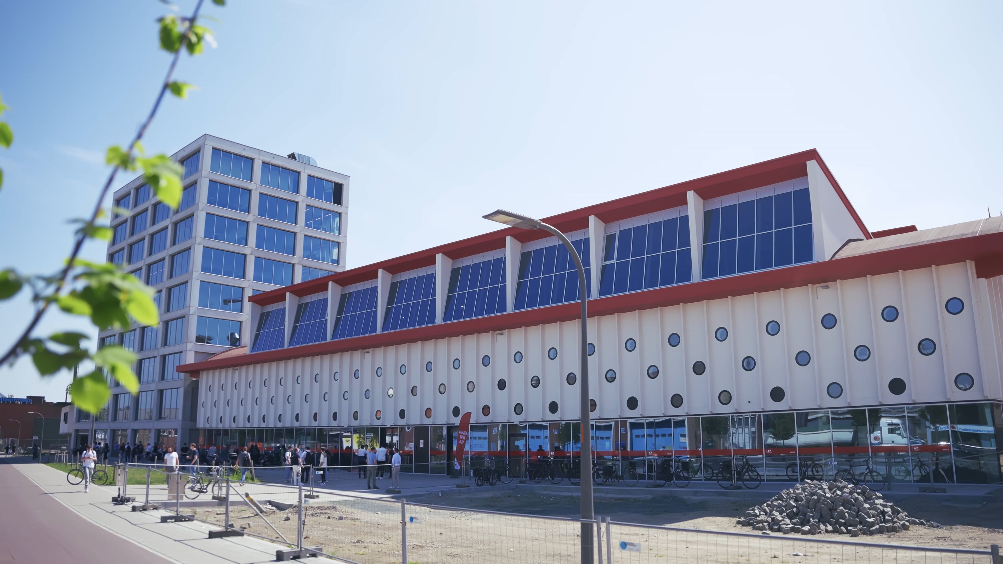 Een modern gebouw met een rood dak en grote blauwe ramen; op een zonnige dag stonden er mensen buiten bijeen.